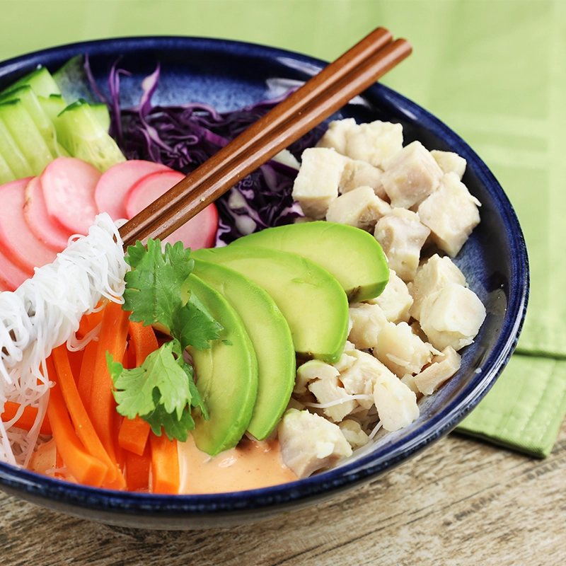 A bowl with diced chicken, avocado, carrots, rice noodles, radish, cucumber, cabbage, and sauce