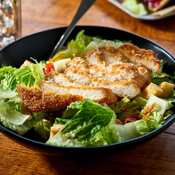Bowl of caesar salad topped with a sliced, breaded chicken filet