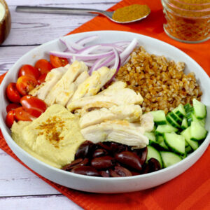 Bowl with sliced chicken, farro, cucumber, olives, hummus, tomatoes, and onions