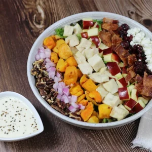 A salad with pecans, onions, squash, chicken, apples, bacon, cranberries, and goat cheese beside of dish of dressing