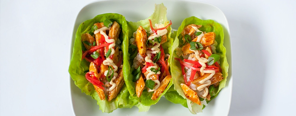 A white plate with three lettuce wraps filled with chicken, veggies, and sauce