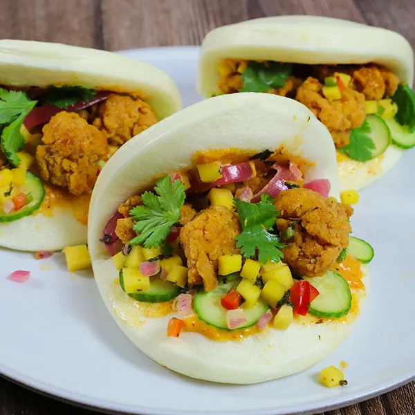 Three bao buns filled with popcorn chicken, lettuce, corn, cilantro, and sauce